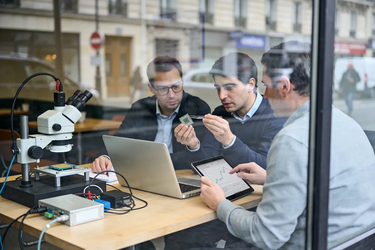 Trois hommes analysent une puce électronique avec laptop et microscope, dans un bureau vitré en ville.