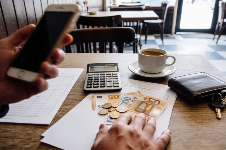 Main tenant téléphone, avec calculatrice, argent, café, clés et portefeuille sur table dans un café.