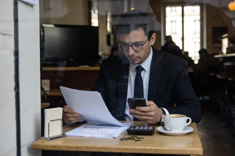 Homme en costume lisant des documents, utilisant un smartphone dans un café, avec un café et calculatrice.