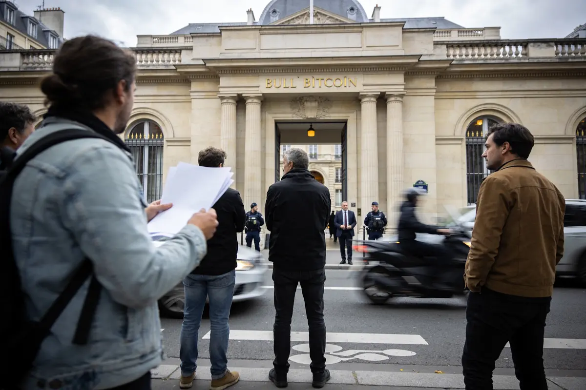 Des gens devant bâtiment "Bull Bitcoin", gardés par policiers, avec scooter passant. Ville en arrière-plan.