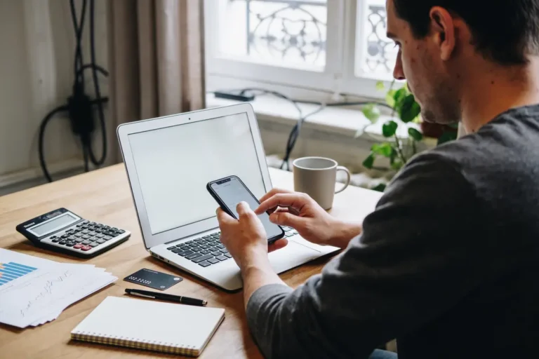 Personne travaillant sur un ordinateur portable avec smartphone, calculatrice, carnet et tasse sur la table.