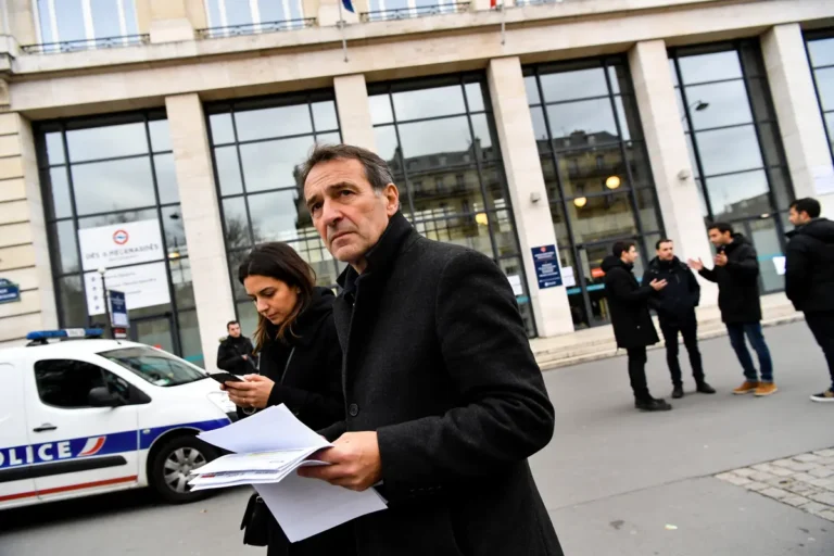 Un homme et une femme, documents en main, devant un bâtiment et une voiture de police.