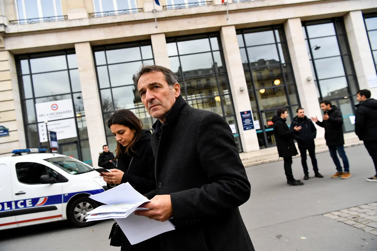 Un homme et une femme, documents en main, devant un bâtiment et une voiture de police.