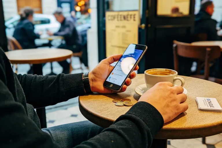 Homme au café consultant smartphone, tasse de café, pièces de monnaie sur table, intérieur de café animé.