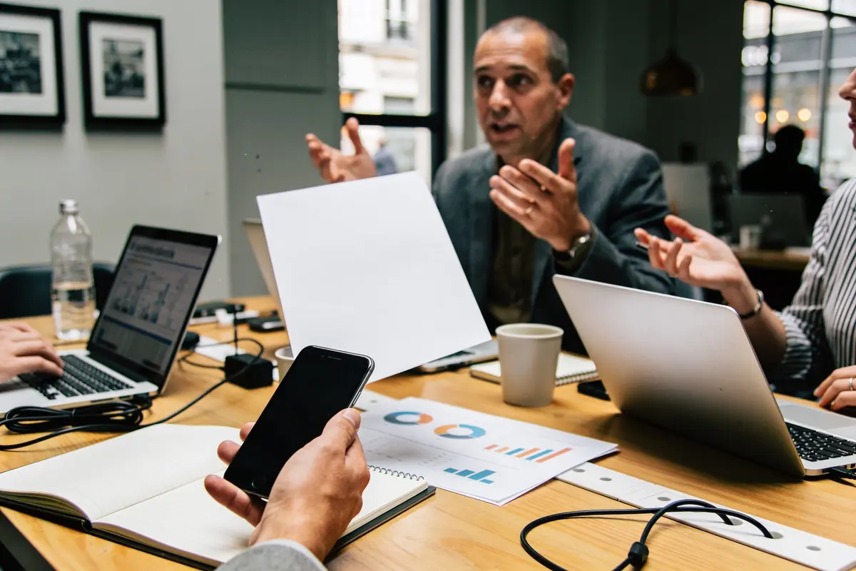 Réunion de travail avec plusieurs personnes discutant autour d'une table, utilisant des ordinateurs portables et des documents.