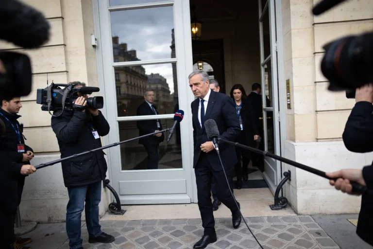 Homme en costume sortant d'un bâtiment, entouré de journalistes et de caméras.