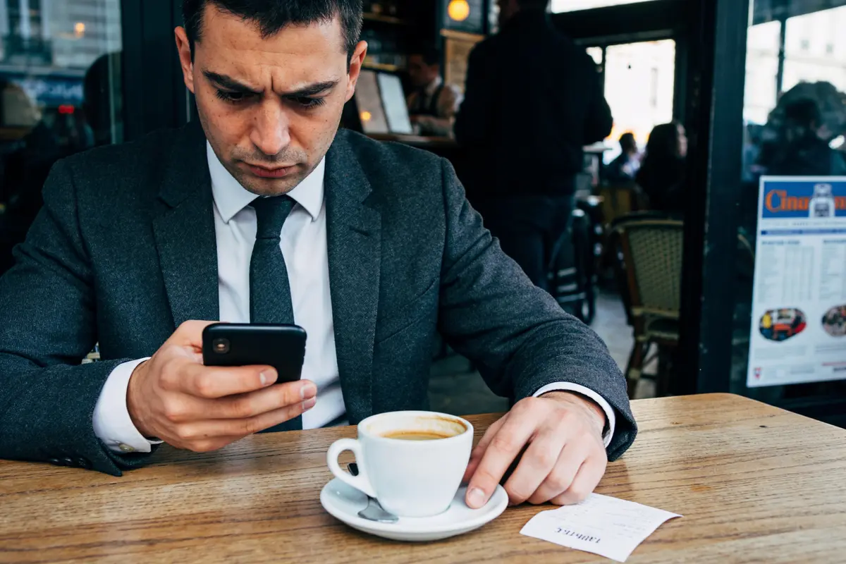 Homme en costume, concentré sur son smartphone, café à la table, menu visible en arrière-plan de café.