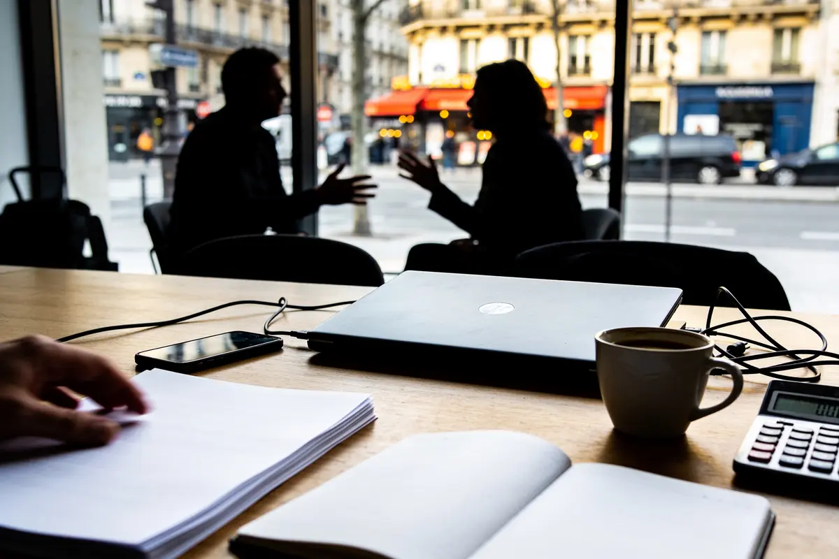 Personnes discutant dans un café, vue à travers la fenêtre. Ordinateur portable, café et documents sur la table.