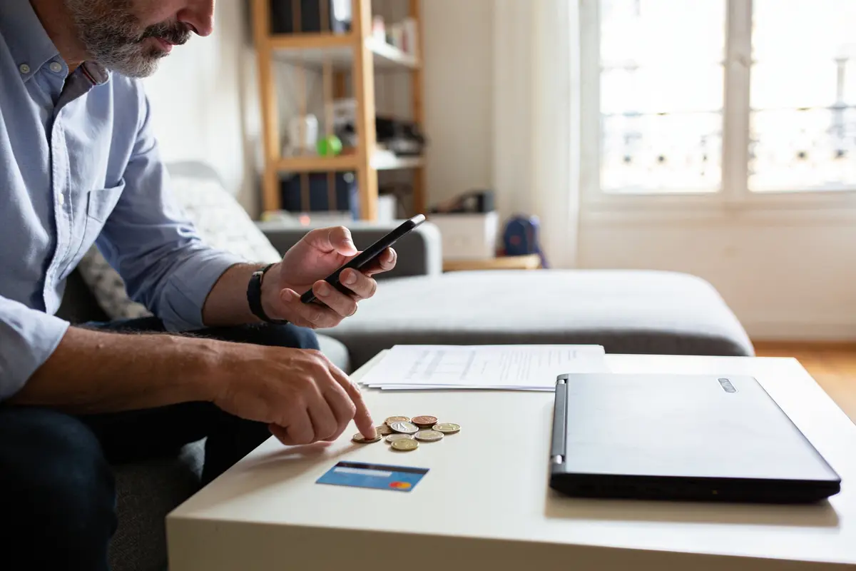 Homme vérifiant téléphone près de pièces, carte bancaire, documents, ordinateur portable sur table basse blanche.