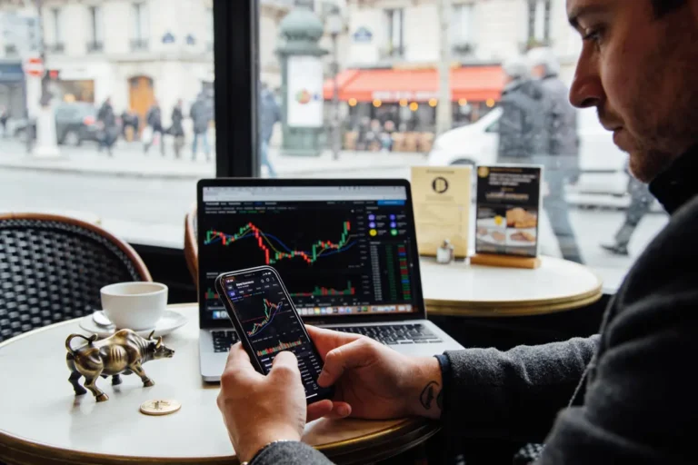 Homme vérifiant cours boursiers sur smartphone et ordinateur depuis un café avec rue visible en arrière-plan.