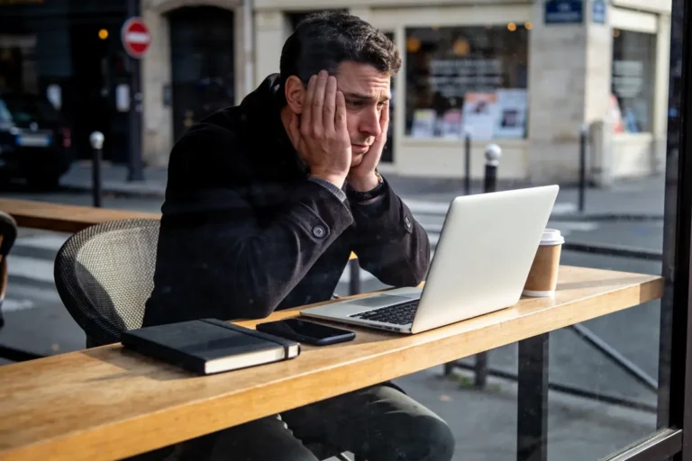 Homme pensif devant ordinateur portable dans café, avec téléphone, carnet et café sur la table.
