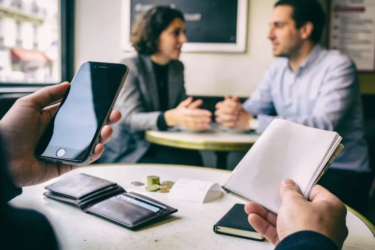 Personnes discutant dans un café, smartphone et carnet au premier plan, avec portefeuille et monnaie sur la table.
