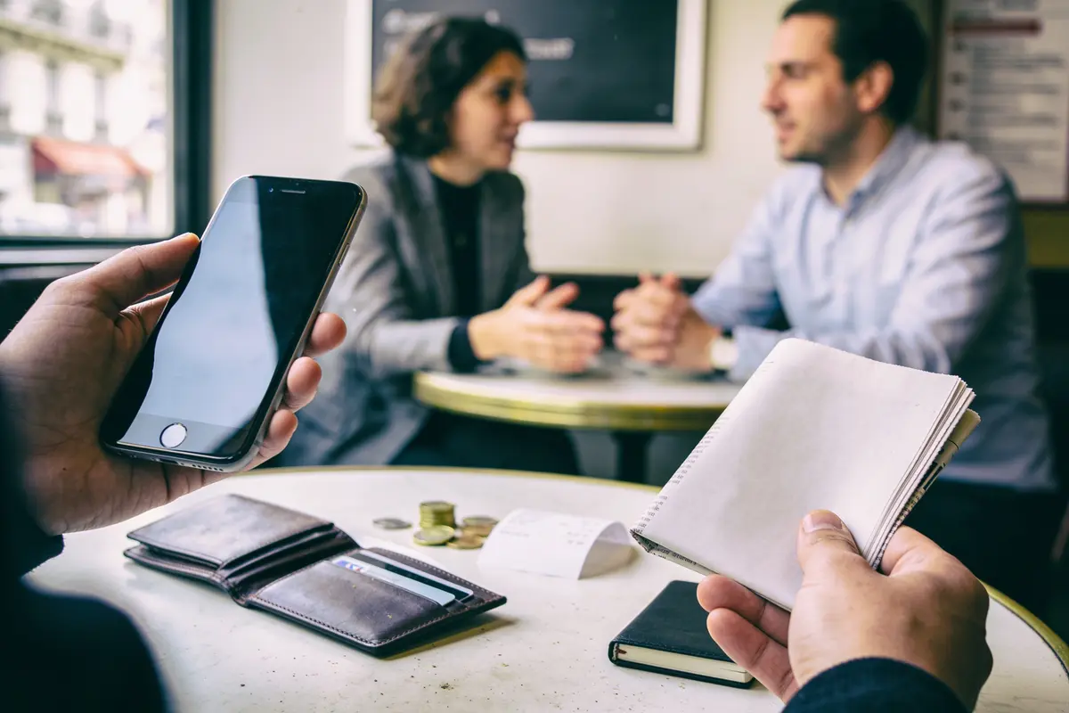 Personnes discutant dans un café, smartphone et carnet au premier plan, avec portefeuille et monnaie sur la table.