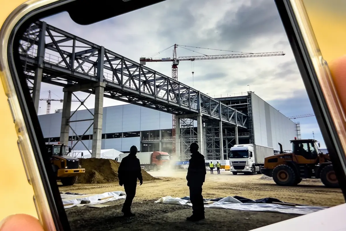 Deux silhouettes sur un chantier de construction avec grue, bâtiment métallique et véhicules lourds autour.
