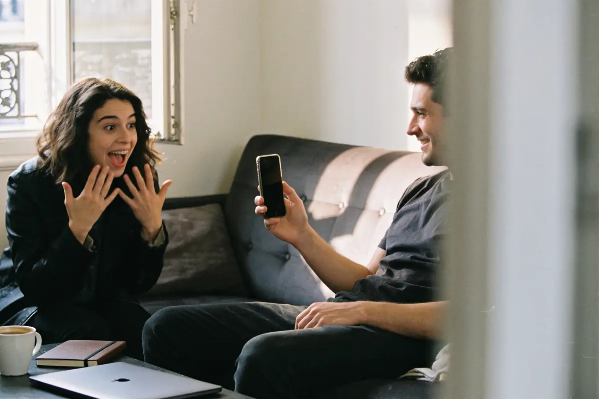 Deux personnes sur un canapé, discutant joyeusement, homme tenant un smartphone, café et ordinateur près d'eux.