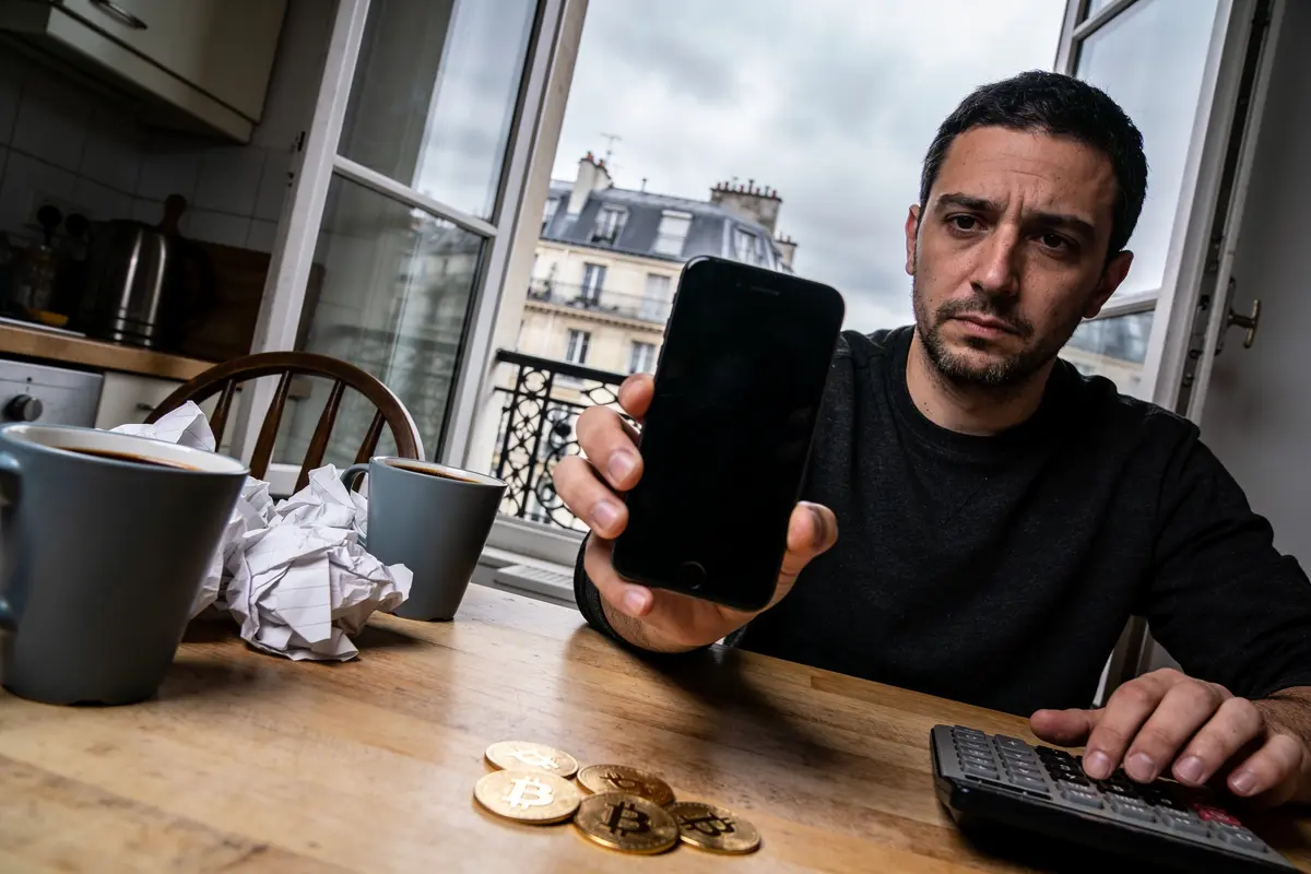 Homme montrant un smartphone éteint, pièces Bitcoin, calculatrice, tasses, fenêtres ouvertes et ciel nuageux.