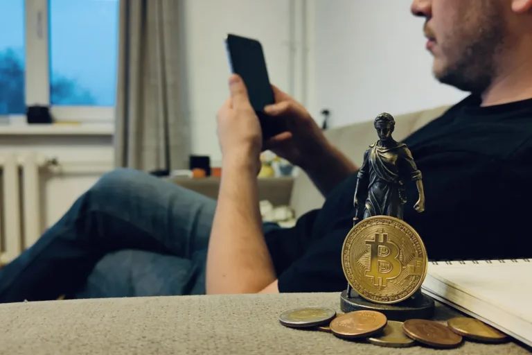 Homme assis sur canapé lisant tablette, statue et pièce Bitcoin en premier plan.
