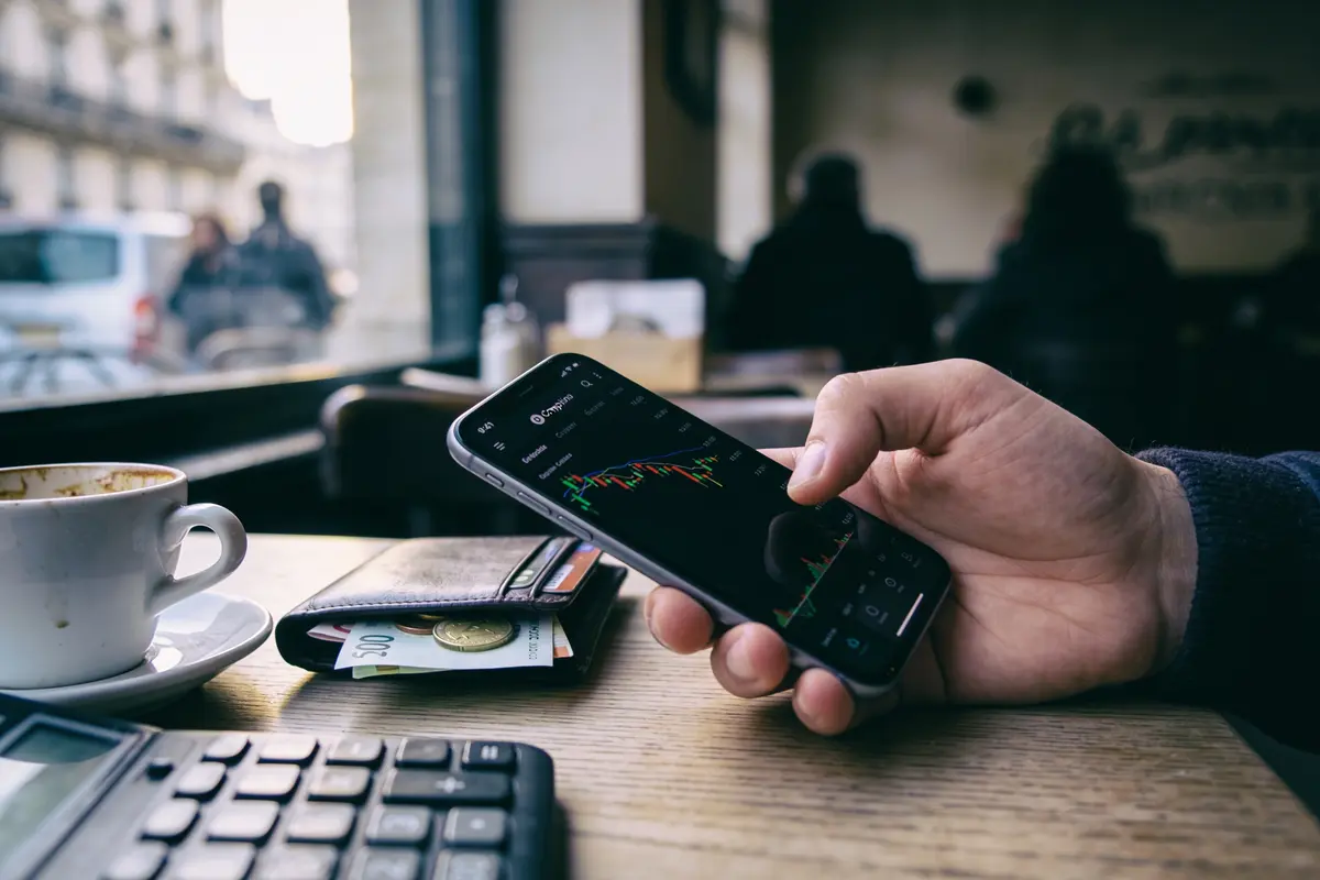 Personne tenant un smartphone affichant un graphique financier, avec une tasse de café et une calculatrice sur la table.