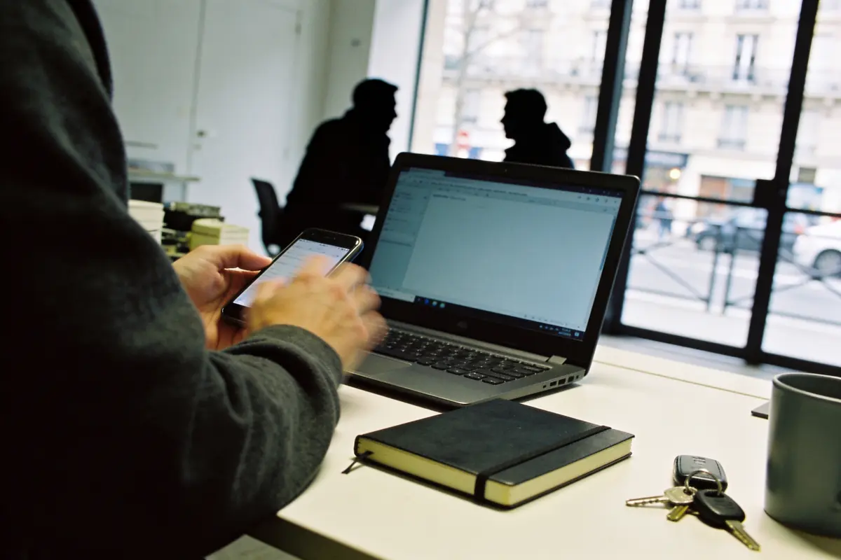 Personne utilisant un smartphone devant un ordinateur portable, carnet noir, clés et tasse sur le bureau.