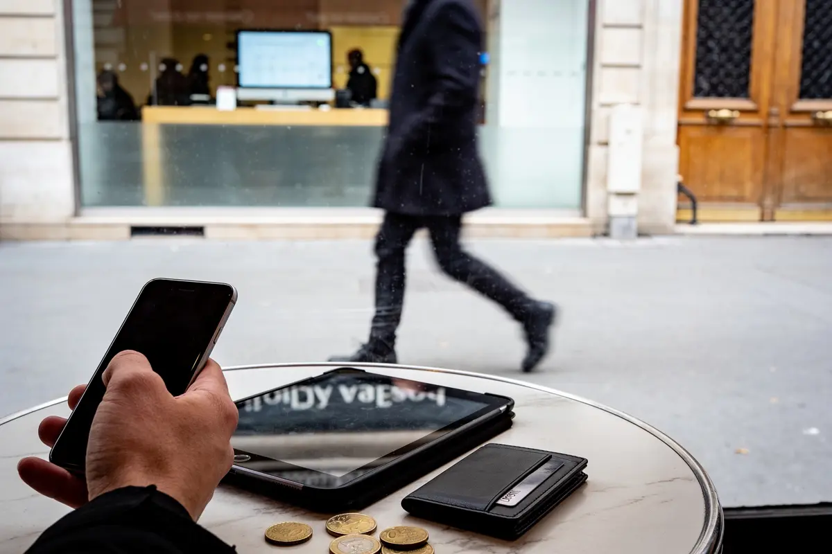 Main tenant un smartphone, pièce, table, homme passant dehors, devant un bâtiment avec écran allumé.