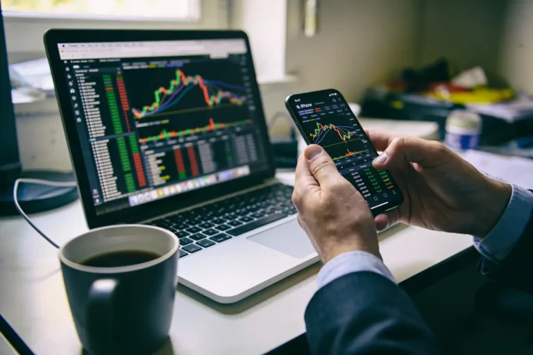 Personne analysant graphiques boursiers sur smartphone et ordinateur portable avec café à proximité.