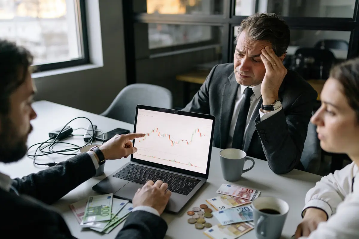Trois personnes discutent d'un graphique financier sur un ordinateur portable, avec des billets euros et pièces.