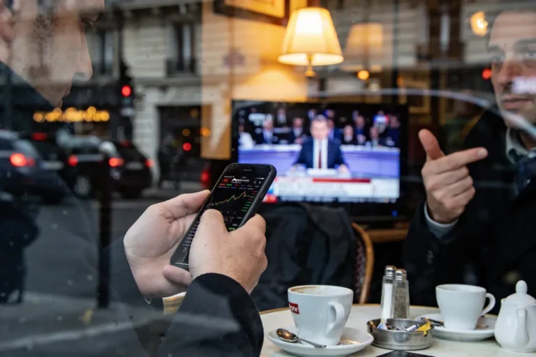 Deux hommes discutent dans un café, l'un regarde son téléphone, télévision en arrière-plan affichant des nouvelles.