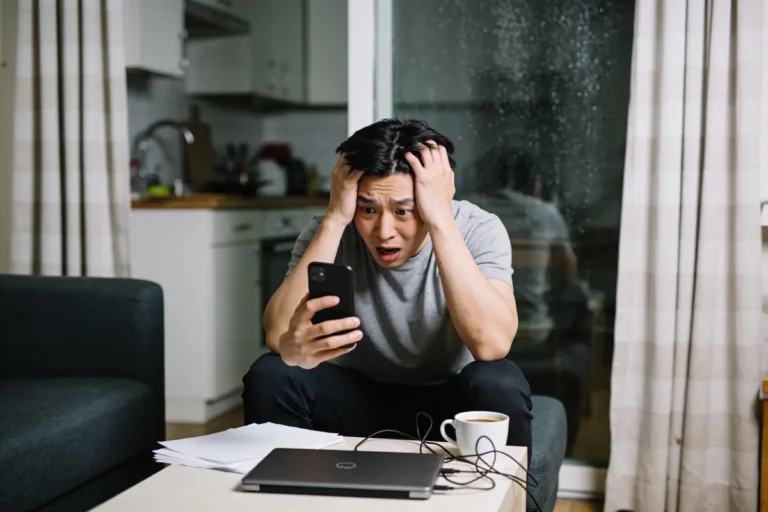 Homme stressé tenant un téléphone, assis, ordinateur portable et café sur la table devant lui.