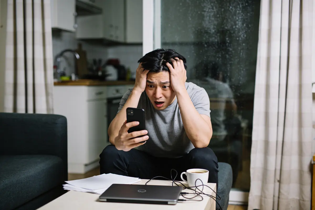 Homme stressé tenant un téléphone, assis, ordinateur portable et café sur la table devant lui.