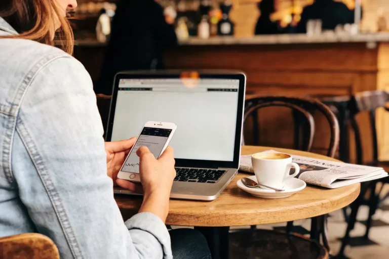 Personne utilisant smartphone et ordinateur portable dans un café, tasse de café et journal sur la table.