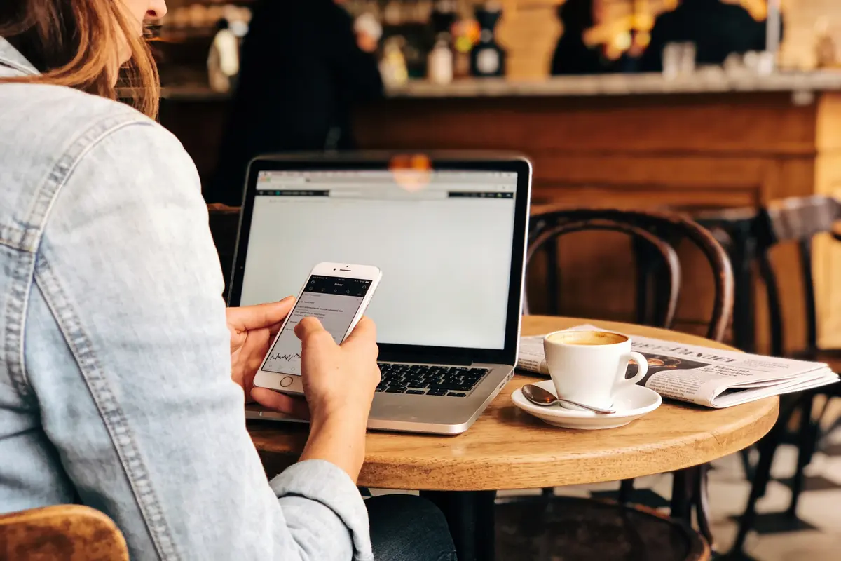 Personne utilisant smartphone et ordinateur portable dans un café, tasse de café et journal sur la table.