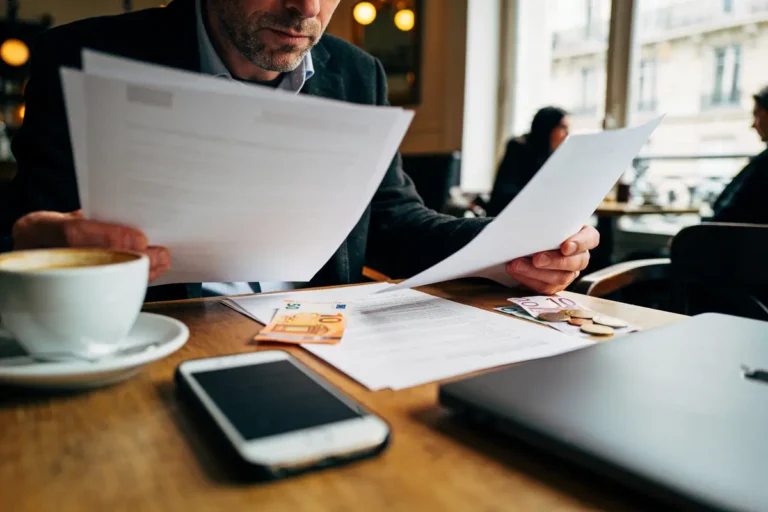 Homme lisant des documents sur une table de café avec smartphone, café, ordinateur portable et euros.