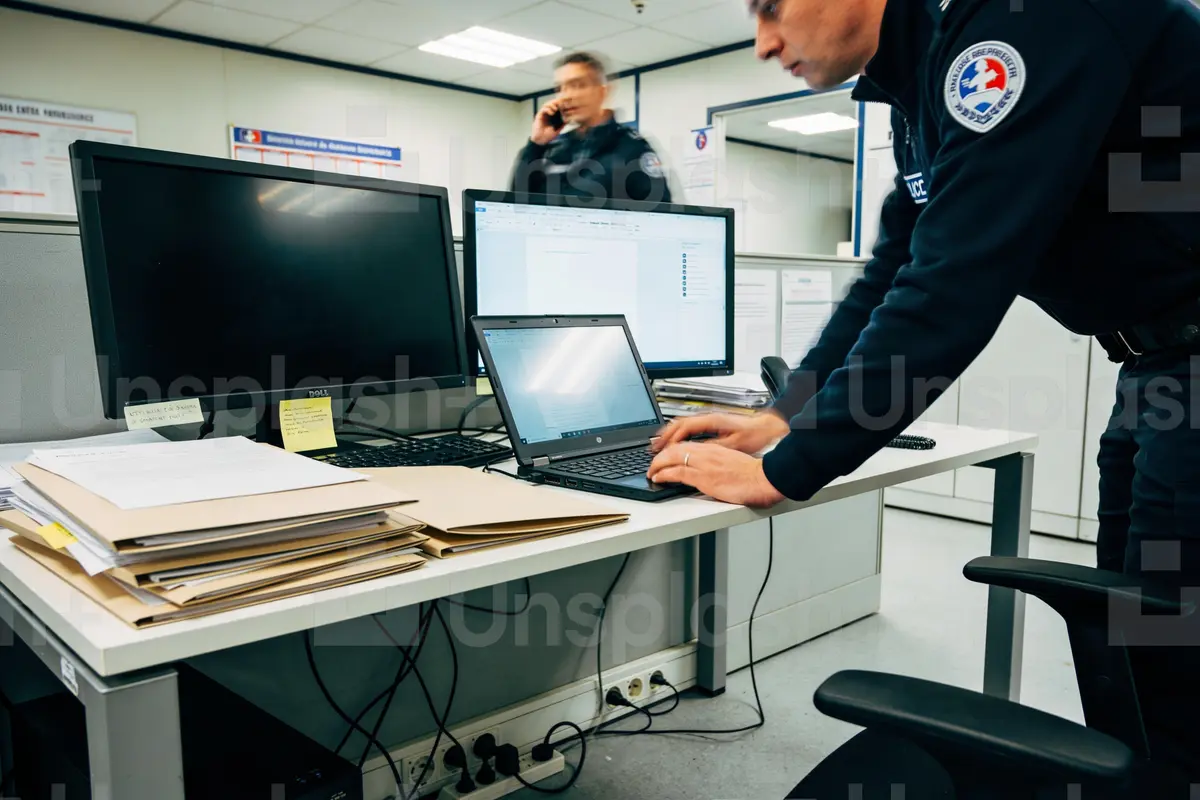 Deux policiers travaillant au bureau, un sur ordinateur portable, l'autre téléphone, dossiers sur le bureau.
