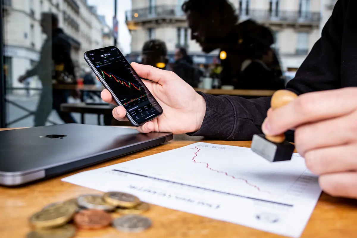 Personne tenant smartphone affichant graphique financier, pièces et ordinateur portable sur table en bois.