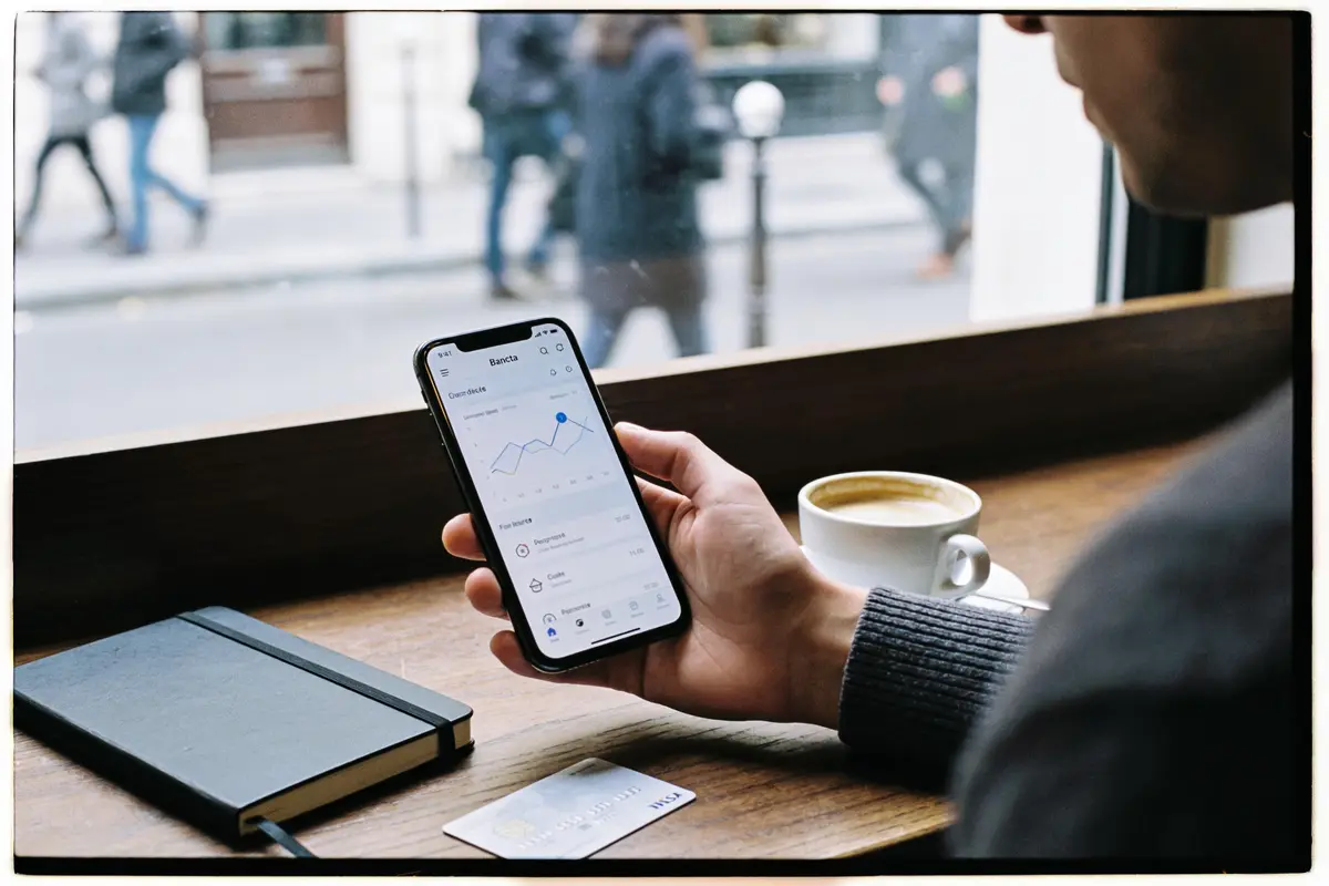 Personne utilisant un smartphone affichant des graphiques, avec un carnet, une carte bancaire et un café posés.