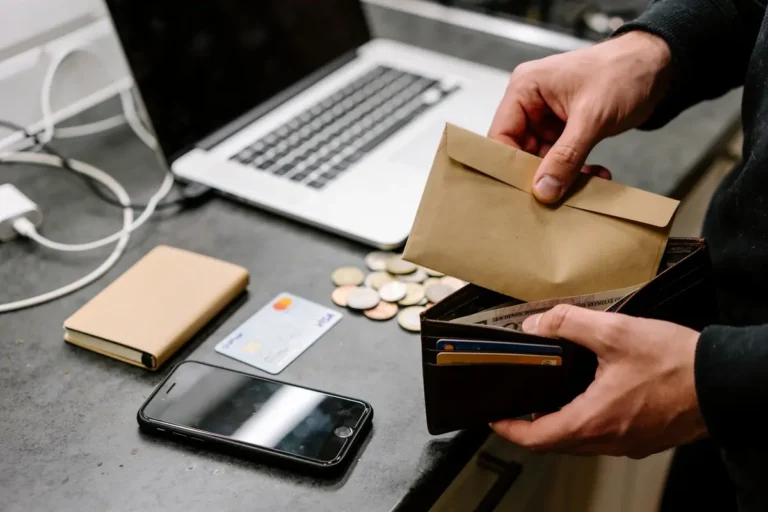 Personne manipulant enveloppe et portefeuille sur table, avec ordinateur, pièce de monnaie, téléphone, et carte bancaire.