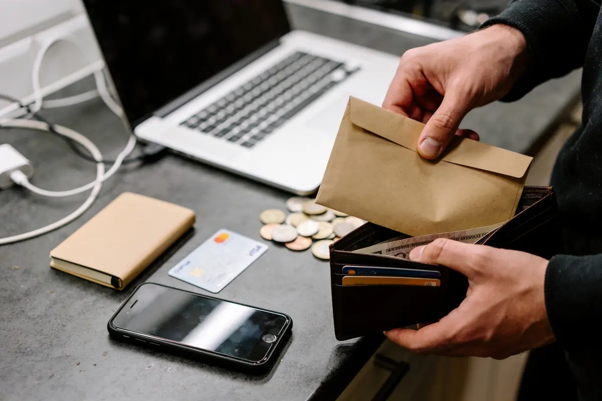 Personne manipulant enveloppe et portefeuille sur table, avec ordinateur, pièce de monnaie, téléphone, et carte bancaire.