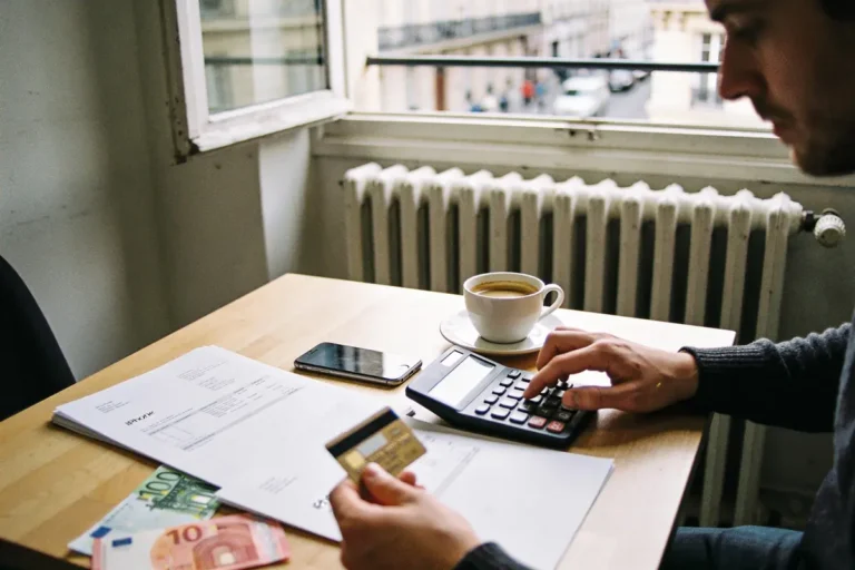 Personne calculant des finances, avec documents, carte bancaire, smartphone, et café sur la table près de la fenêtre.