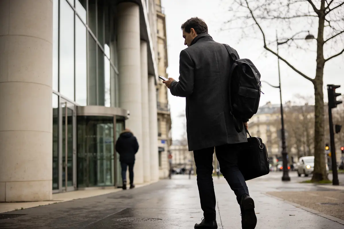 Homme en manteau sombre marche sur trottoir, regardant son téléphone, devant un bâtiment moderne vitré.