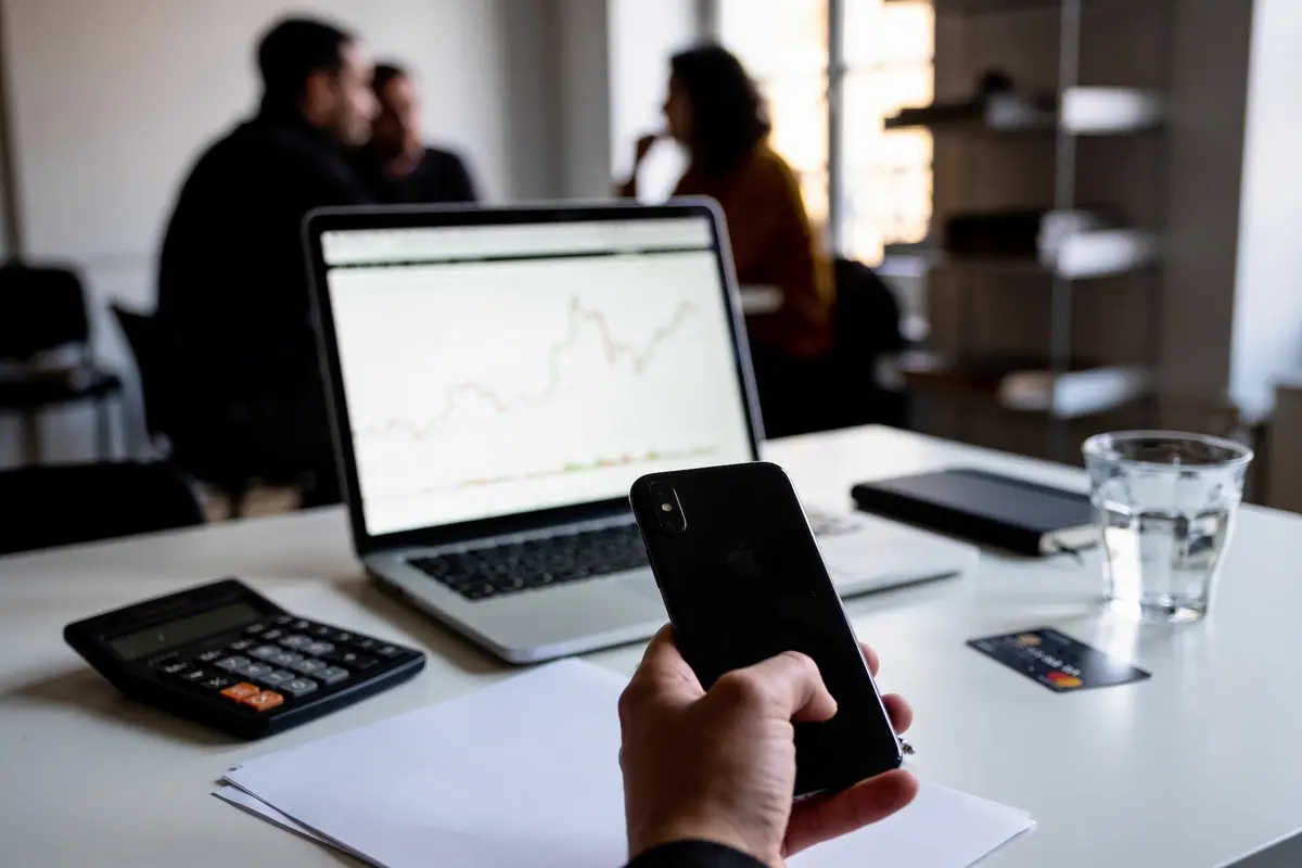 Personne tenant un smartphone devant un ordinateur affichant un graphique financier, avec une calculette et un verre.