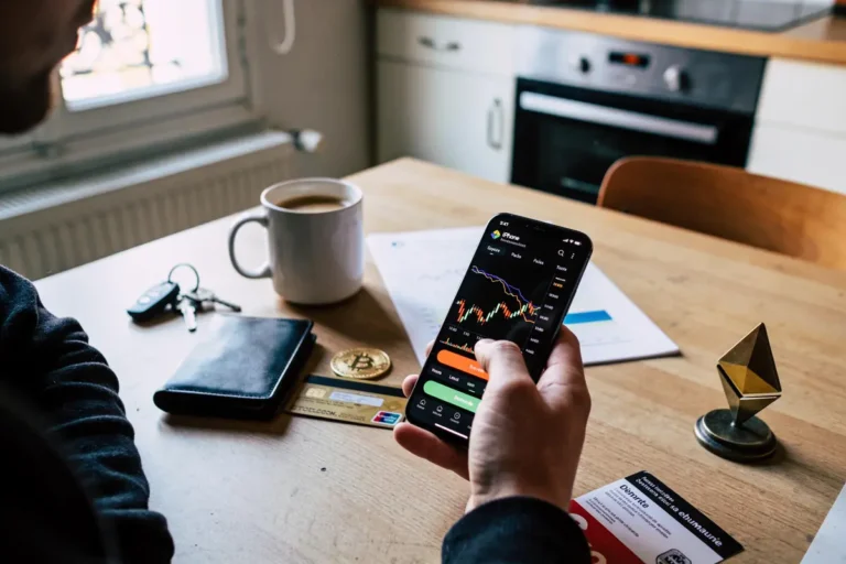 Personne utilisant smartphone avec graphiques financiers, table avec portefeuille, clés, tasse café, cartes et crypto-monnaie.