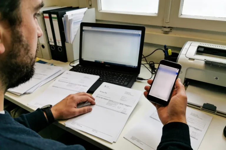 Un homme travaille à un bureau avec ordinateur portable, documents, smartphone et imprimante.