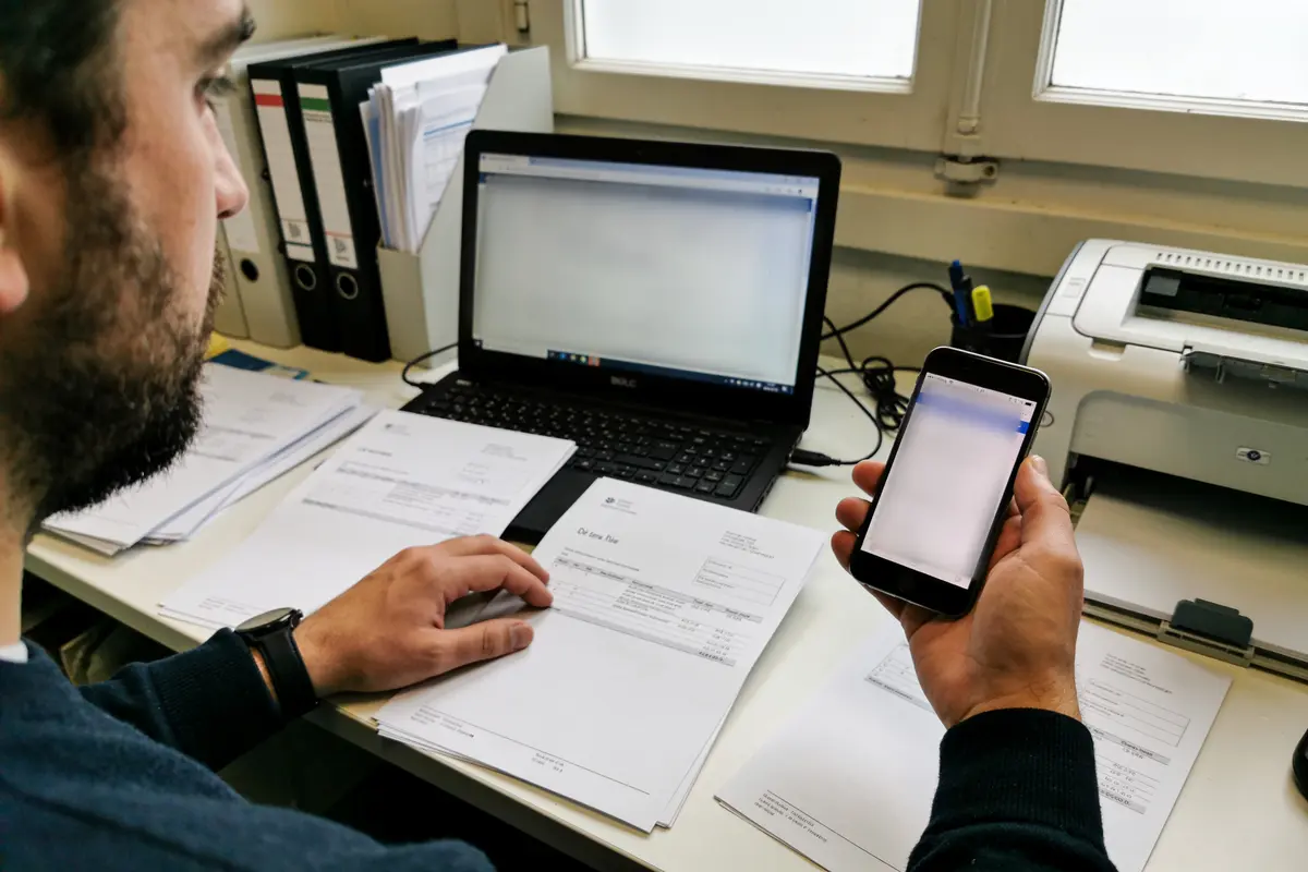 Un homme travaille à un bureau avec ordinateur portable, documents, smartphone et imprimante.