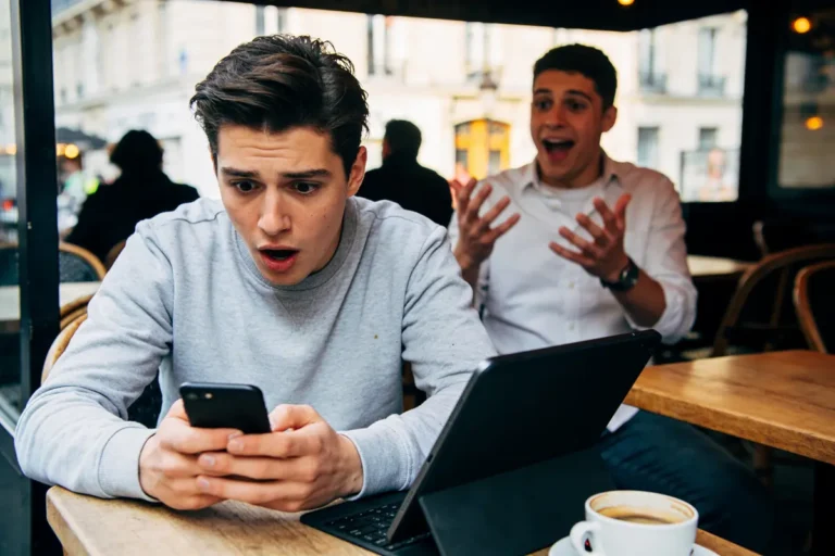 Deux hommes surpris, assis dans un café, regardent un téléphone et un ordinateur portable.