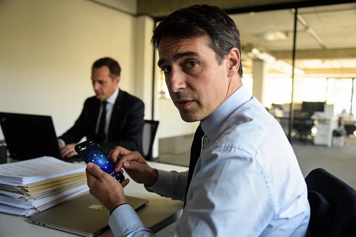 Homme en chemise blanche tenant un smartphone, collègue en costume travaillant sur ordinateur portable. Bureau lumineux.
