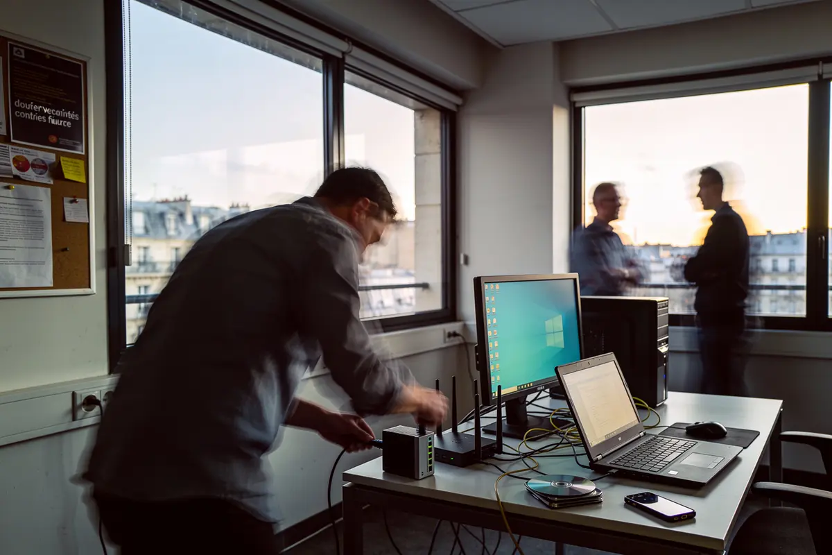 Homme ajustant équipement informatique, discussions en arrière-plan, bureaux modernes, lumière du jour à travers les fenêtres.