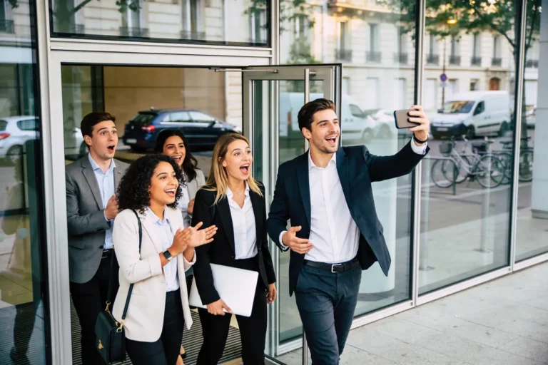 Groupe de collègues souriants prenant un selfie devant un bâtiment avec des voitures et vélos en arrière-plan.