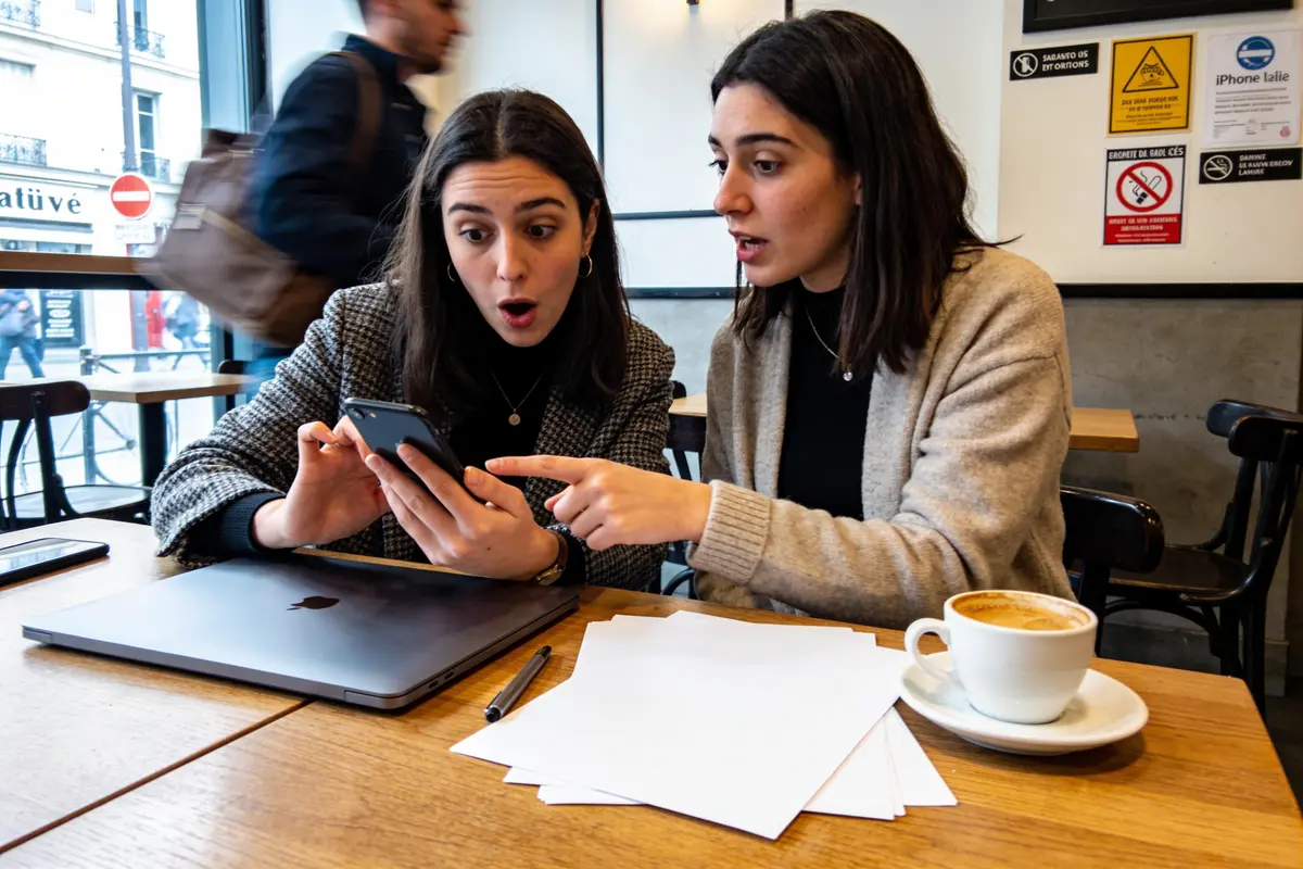 Deux femmes surprises regardent un smartphone dans un café, avec des documents et un ordinateur portable.