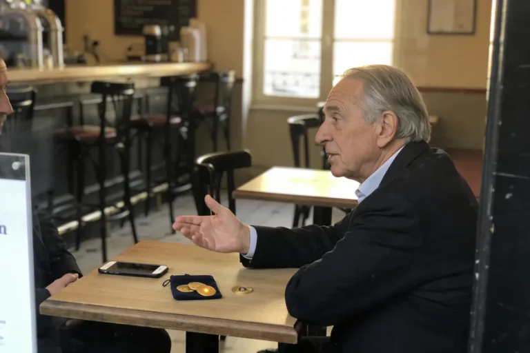 Un homme discute dans un café avec téléphone et pièces en or sur la table.