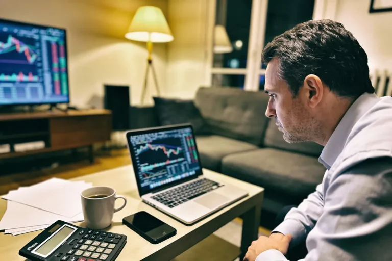 Homme analysant graphiques financier sur ordinateur portable, environnement moderne, avec tasse de café et calculatrice.